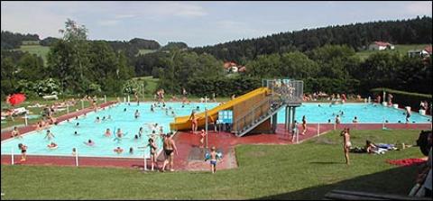 Öffentliches Freibad mit Menschen, die schwimmen und sich entspannen; eine gelbe Rutsche teilt das Becken, mit Grasflächen und Bäumen im Hintergrund.