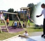 Kinder spielen auf einem hölzernen Spielplatz mit Schaukeln und Klettergerüsten, während ein anderes Kind auf einer gepflasterten Fläche in einem Park im Freien Rollschuh fährt.