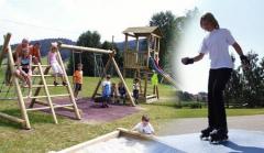 Kinder spielen auf einem hölzernen Spielplatz mit Schaukeln und Klettergerüsten, während ein anderes Kind auf einer gepflasterten Fläche in einem Park im Freien Rollschuh fährt.