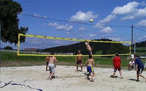 Sechs Personen spielen Beachvolleyball auf einem Sandplatz im Freien; ein Spieler springt, um den Ball bei teilweise bewölktem Himmel über das Netz zu schlagen.