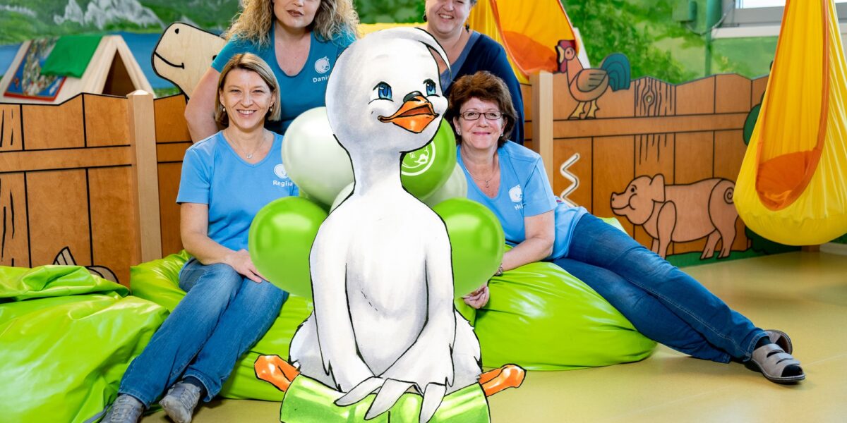 Vier Erwachsene in blauen Hemden sitzen auf grünen Sitzsäcken in einem farbenfrohen Raum, in dem eine große illustrierte Entenfigur im Vordergrund steht.