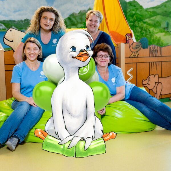 Vier Erwachsene in blauen Hemden sitzen auf grünen Sitzsäcken in einem farbenfrohen Raum, in dem eine große illustrierte Entenfigur im Vordergrund steht.