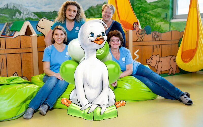 Vier Erwachsene in blauen Hemden sitzen auf grünen Sitzsäcken in einem farbenfrohen Raum, in dem eine große illustrierte Entenfigur im Vordergrund steht.