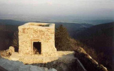 Ein kleines, verwittertes Steingebäude mit einer Tür steht an einem Berghang und überblickt eine ferne Landschaft unter einem dunstigen Himmel.