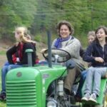 Vier Personen fahren auf einem grünen Traktor in einem grasbewachsenen Feld mit Bäumen im Hintergrund.