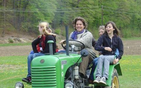 Vier Personen fahren auf einem grünen Traktor in einem grasbewachsenen Feld mit Bäumen im Hintergrund.