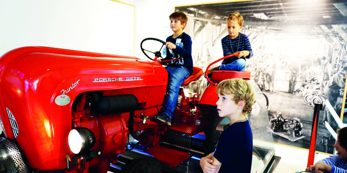 Drei Jungen interagieren mit einem alten roten Porsche Diesel Junior Traktor, der in einer Halle ausgestellt ist; zwei sitzen auf dem Traktor, während einer in der Nähe steht.