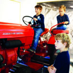 Drei Jungen interagieren mit einem alten roten Porsche Diesel Junior Traktor, der in einer Halle ausgestellt ist; zwei sitzen auf dem Traktor, während einer in der Nähe steht.