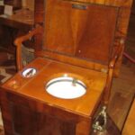 Ein alter hölzerner Toilettenstuhl mit einem geöffneten Klappdeckel, der einen Nachttopf aus Keramik enthüllt.