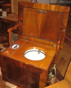 Ein alter hölzerner Toilettenstuhl mit einem geöffneten Klappdeckel, der einen Nachttopf aus Keramik enthüllt.