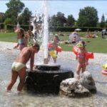 Kinder in Badeanzügen und mit Schwimmflügeln spielen in einem flachen Becken in einem Park um eine Wasserfontäne herum, während im Hintergrund Menschen auf dem Rasen faulenzen.