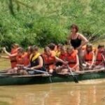 Eine Gruppe von Kindern und ein Erwachsener, die Schwimmwesten tragen, paddeln in einem Kanu einen schlammigen Fluss hinunter, der von Grünflächen umgeben ist.