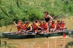 Eine Gruppe von Kindern und ein Erwachsener, die Schwimmwesten tragen, paddeln in einem Kanu einen schlammigen Fluss hinunter, der von Grünflächen umgeben ist.