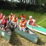 Eine Gruppe von Personen mit Schwimmwesten sitzt in Kanus am Ufer und bereitet sich darauf vor, auf einem Fluss zu paddeln, mit Bäumen und Gras im Hintergrund.