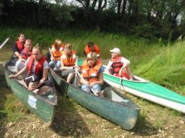 Eine Gruppe von Personen mit Schwimmwesten sitzt in Kanus am Ufer und bereitet sich darauf vor, auf einem Fluss zu paddeln, mit Bäumen und Gras im Hintergrund.
