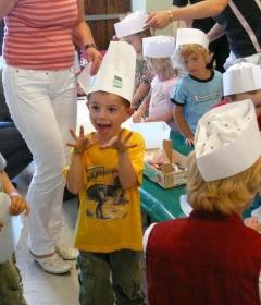 Eine Gruppe von Kindern mit Kochmützen aus Papier nimmt an einer Aktivität teil; ein Kind in einem gelben Hemd steht vorne mit erhobenen Händen und scheint aufgeregt zu sein.