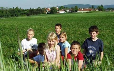 Eine Gruppe von acht Kindern sitzt und kniet auf einer grasbewachsenen Wiese mit einem Dorf und Hügeln im Hintergrund.