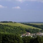 Ein kleines Dorf mit einer Kirche liegt eingebettet zwischen grünen Hügeln und Wäldern unter einem teilweise bewölkten Himmel.