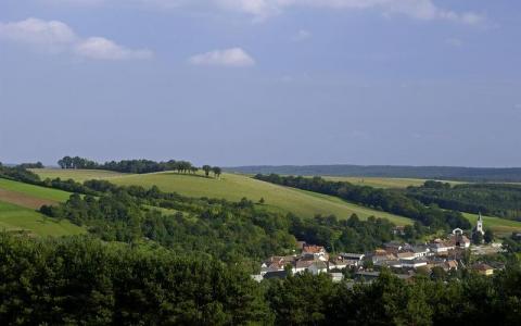 Ein kleines Dorf mit einer Kirche liegt eingebettet zwischen grünen Hügeln und Wäldern unter einem teilweise bewölkten Himmel.