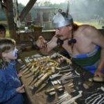 Ein Mann mit Hornhelm zeigt einem Kind mit Gesichtsbemalung bei einer historischen Nachstellung im Freien Knochenwerkzeuge und Schmuck.
