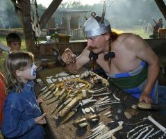Ein Mann mit Hornhelm zeigt einem Kind mit Gesichtsbemalung bei einer historischen Nachstellung im Freien Knochenwerkzeuge und Schmuck.
