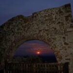 Im Vordergrund steht ein steinerner Torbogen, durch den ein Sonnenuntergang vor einem sich verdunkelnden Himmel zu sehen ist.