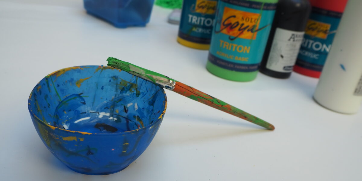 Eine blaue Farbschale mit einem grünen Pinsel auf dem Rand steht auf einem Tisch, umgeben von Flaschen mit Acrylfarben und Kunstzubehör.