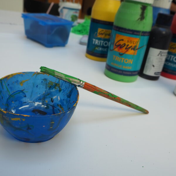 Eine blaue Farbschale mit einem grünen Pinsel auf dem Rand steht auf einem Tisch, umgeben von Flaschen mit Acrylfarben und Kunstzubehör.
