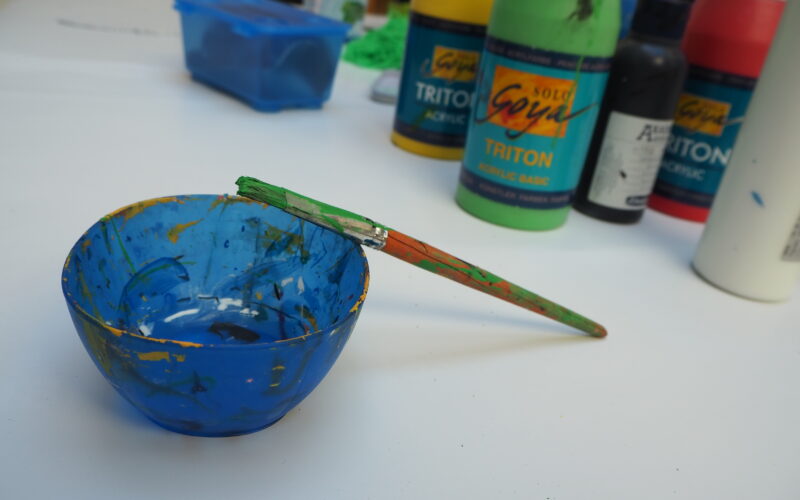 Eine blaue Farbschale mit einem grünen Pinsel auf dem Rand steht auf einem Tisch, umgeben von Flaschen mit Acrylfarben und Kunstzubehör.