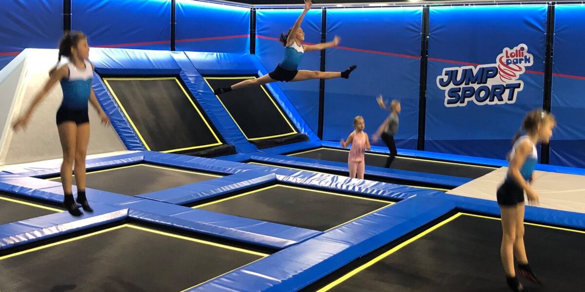 Fünf Kinder springen auf Trampolinen in einem Indoor-Trampolinpark namens "Jump Sport". Ein Kind führt einen Spagatsprung in der Luft aus.