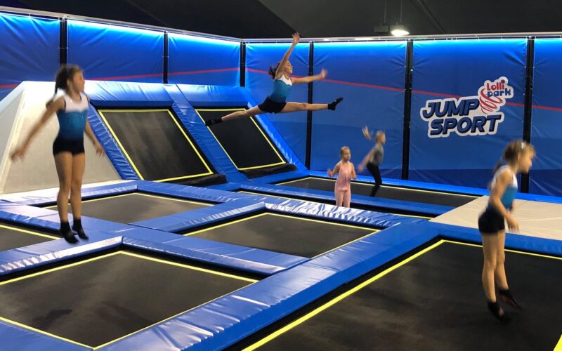 Fünf Kinder springen auf Trampolinen in einem Indoor-Trampolinpark namens "Jump Sport". Ein Kind führt einen Spagatsprung in der Luft aus.