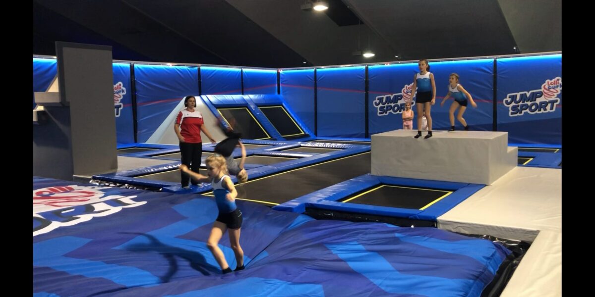 Kinder und ein Erwachsener befinden sich in einem Indoor-Trampolinpark mit blauen Matten, Trampolinen und dem Jump-Sport-Logo an den Wänden.