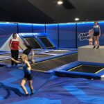 Kinder und ein Erwachsener befinden sich in einem Indoor-Trampolinpark mit blauen Matten, Trampolinen und dem Jump-Sport-Logo an den Wänden.