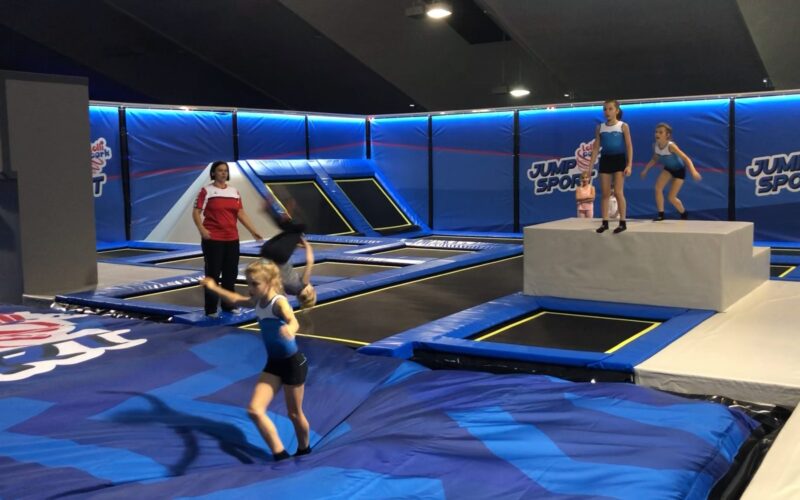 Kinder und ein Erwachsener befinden sich in einem Indoor-Trampolinpark mit blauen Matten, Trampolinen und dem Jump-Sport-Logo an den Wänden.