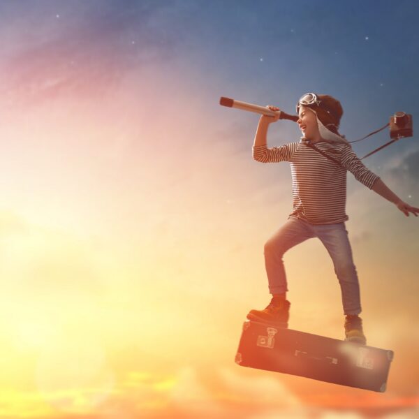 Ein Kind mit Fliegerbrille steht auf einem schwimmenden Koffer und blickt durch ein Teleskop mit einer Kamera um den Hals in den Sonnenuntergangshimmel.