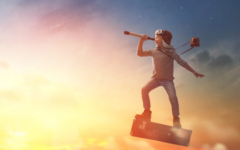 Ein Kind mit Fliegerbrille steht auf einem schwimmenden Koffer und blickt durch ein Teleskop mit einer Kamera um den Hals in den Sonnenuntergangshimmel.