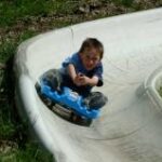 Ein Kind fährt an einem sonnigen Tag im Freien auf einem blauen Schlitten eine Betonrutsche hinunter.