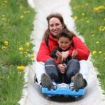 Ein Erwachsener und ein Kind fahren gemeinsam auf einem blauen Schlitten eine Betonrutsche hinunter, umgeben von Gras und gelben Blumen.