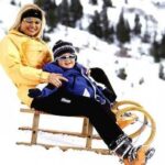 Ein Erwachsener und ein Kind in Winterkleidung sitzen zusammen auf einem Holzschlitten in einer verschneiten Umgebung mit Bäumen im Hintergrund.