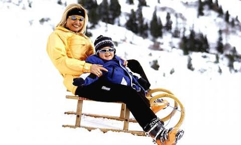 Ein Erwachsener und ein Kind in Winterkleidung sitzen zusammen auf einem Holzschlitten in einer verschneiten Umgebung mit Bäumen im Hintergrund.