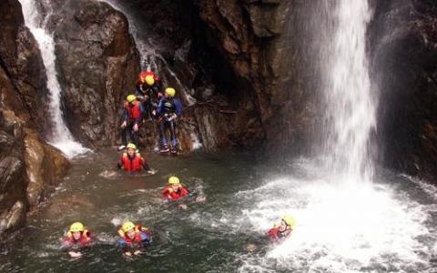 Eine Gruppe von Menschen mit Helmen und Schwimmwesten schwimmt und steht in einem Felsbecken unter einem Wasserfall.