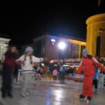 Kinder, die nachts im Freien Schlittschuh laufen, mit beleuchteten Gebäuden im Hintergrund.