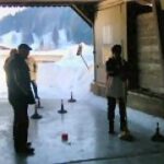 Mehrere Personen stehen auf einer Eisbahn in einer Halle und spielen ein Spiel mit Besenstielen und Metallgewichten. Außerhalb der offenen Struktur ist eine verschneite Landschaft zu sehen.