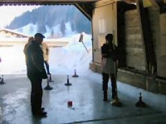 Mehrere Personen stehen auf einer Eisbahn in einer Halle und spielen ein Spiel mit Besenstielen und Metallgewichten. Außerhalb der offenen Struktur ist eine verschneite Landschaft zu sehen.