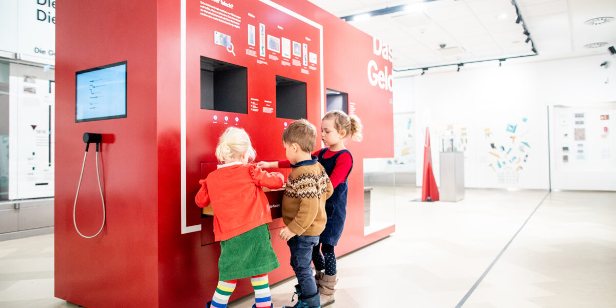 Drei kleine Kinder interagieren mit einem großen roten Touchscreen-Kiosk in einem hellen, modernen Ausstellungsraum.