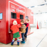 Drei kleine Kinder interagieren mit einem großen roten Touchscreen-Kiosk in einem hellen, modernen Ausstellungsraum.