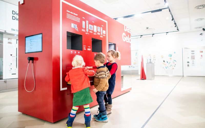 Drei kleine Kinder interagieren mit einem großen roten Touchscreen-Kiosk in einem hellen, modernen Ausstellungsraum.