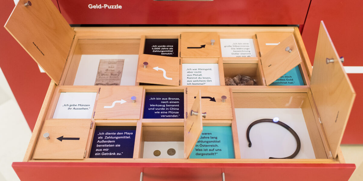Eine offene Schublade mit der Aufschrift "Setz-Kasten" enthält unterteilte Fächer mit Münzen, Textkarten, Fotos und einem schwarzen Armband, das Teil eines Displays mit der Aufschrift "Geld-Puzzle" ist.