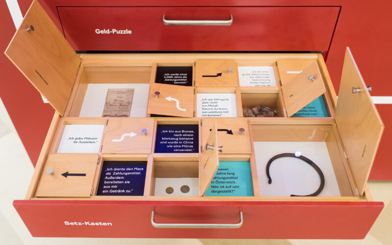 Eine offene Schublade mit der Aufschrift "Setz-Kasten" enthält unterteilte Fächer mit Münzen, Textkarten, Fotos und einem schwarzen Armband, das Teil eines Displays mit der Aufschrift "Geld-Puzzle" ist.