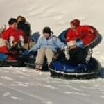 Mehrere Personen, darunter Kinder und Erwachsene, sitzen in Snowtubes und rutschen gemeinsam einen verschneiten Hügel hinunter.